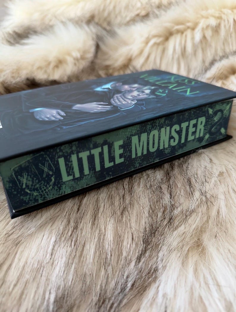 Pu&ograve; includere: Un libro con copertina rigida con una copertina scura con l'illustrazione di due figure. Il dorso del libro mostra il testo "LITTLE MONSTER" in lettere verdi. Il libro &egrave; appoggiato su una superficie soffice e chiara.