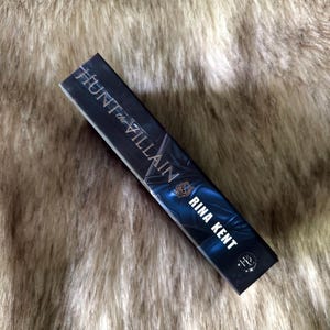 K&ouml;nnte beinhalten: Ein Buch mit dem Titel "Hunt the Villain" von Rina Kent. Das Buch hat einen dunklen Einband mit blauen und silbernen Akzenten. Der Name des Autors ist in Wei&szlig; gehalten. Das Buch ist rechteckig und scheint ein Taschenbuch zu sein.
