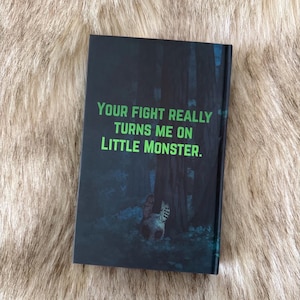 Pu&ograve; includere: Un libro con una copertina scura con il testo verde "YOUR FIGHT REALLY TURNS ME ON LITTLE MONSTER". Lo sfondo raffigura una scena di foresta con un gatto. Il libro &egrave; appoggiato su una superficie pelosa.