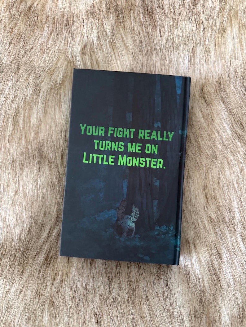 Pu&ograve; includere: Un libro con una copertina scura con il testo verde "YOUR FIGHT REALLY TURNS ME ON LITTLE MONSTER". Lo sfondo raffigura una scena di foresta con un gatto. Il libro &egrave; appoggiato su una superficie pelosa.