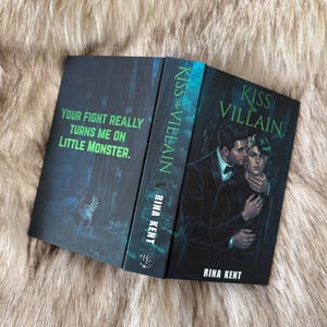 Pu&ograve; includere: Libro aperto intitolato "Kiss the Villain" di Rina Kent. La copertina presenta una coppia che si abbraccia, con toni verde scuro e nero. La pagina sinistra aperta recita "Your fight really turns me on little monster."