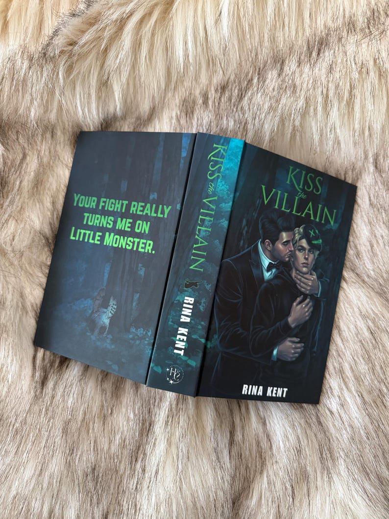 Pu&ograve; includere: Libro aperto intitolato "Kiss the Villain" di Rina Kent. La copertina presenta una coppia che si abbraccia, con toni verde scuro e nero. La pagina sinistra aperta recita "Your fight really turns me on little monster."