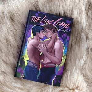 May include: A book titled "THE LONG GAME" with an illustration of two shirtless men embracing. The cover art features a dark background with purple and green accents. The men are wearing blue trousers and red belts.