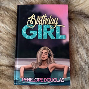 May include: A book cover with the title "Birthday Girl" in gold script and turquoise block letters. The cover features an illustration of a woman in a pool, with the author's name, Penelope Douglas, at the bottom. The background is a gradient of pink and purple.