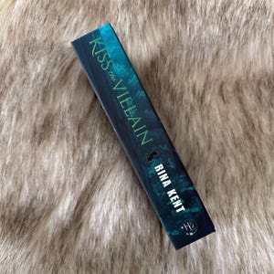 Pu&ograve; includere: Un libro con il titolo "Kiss the Villain" di Rina Kent. La copertina &egrave; verde scuro con testo bianco. Il libro &egrave; appoggiato su una superficie morbida e strutturata di colore marrone chiaro. Il titolo e il nome dell'autore sono bianchi.