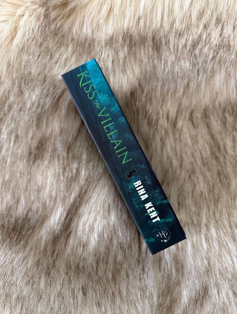 Pu&ograve; includere: Un libro con il titolo "Kiss the Villain" di Rina Kent. La copertina &egrave; verde scuro con testo bianco. Il libro &egrave; appoggiato su una superficie morbida e strutturata di colore marrone chiaro. Il titolo e il nome dell'autore sono bianchi.