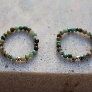 Op de afbeelding: Twee kralenringen met kleine, gefacetteerde turquoise en bruine stenen kralen. De ene ring bevat een enkele gepolijste goudkleurige kraal, terwijl de andere ring een enkele gepolijste zilverkleurige kraal heeft, beide in een cirkelvormig ontwerp.