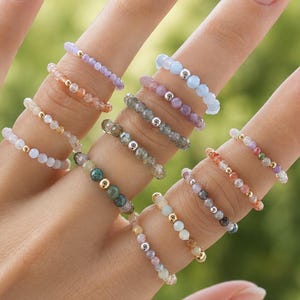 Op de afbeelding: Een collectie delicate elastische ringen gemaakt van kleine, veelkleurige gefacetteerde edelsteenkralen. Elke ring is voorzien van een enkele metalen accentkraal en toont een verscheidenheid aan tinten, waaronder zachtroze, blauw, groen, grijs en perzik.