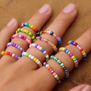 Può includere: Una collezione di dieci anelli realizzati con piccole perline di vetro colorate. Ogni anello presenta un motivo unico con tonalità vivaci, tra cui rosso, arancione, giallo, verde, blu, viola e rosa, disposte in fasce circolari per le dita.