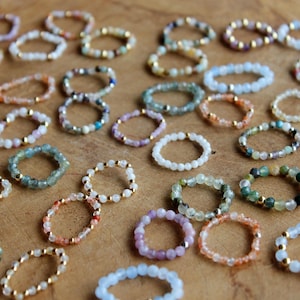 May include: A collection of numerous beaded rings arranged in a scattered pattern. Each ring is constructed from small, multi-colored gemstone beads interspersed with tiny, reflective gold-toned metal spacer beads throughout the circular bands.