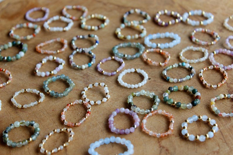 May include: A collection of numerous beaded rings arranged in a scattered pattern. Each ring is constructed from small, multi-colored gemstone beads interspersed with tiny, reflective gold-toned metal spacer beads throughout the circular bands.