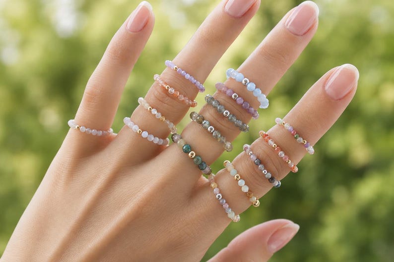 May include: A collection of elastic rings featuring small, multi-colored faceted gemstone beads and tiny metallic gold or silver spacer beads. The rings are worn on multiple fingers, showcasing various hues including light blue, pink, green, and clear.