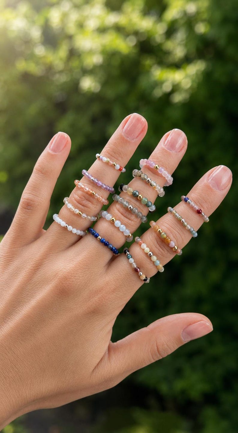 May include: A collection of elastic rings made from small, multicolored gemstone beads and metallic accents. Each ring features a unique combination of translucent, opaque, and metallic spherical beads arranged in a circular band design.