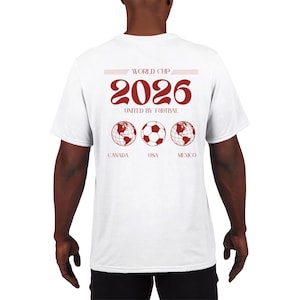 Könnte beinhalten: Ein weißes T-Shirt mit einer roten Grafik auf der Rückseite. Das Design enthält den Text WORLD CUP 2026 UNITED BY FOOTBAL, zwei Globus-Illustrationen, einen Fußball sowie die Beschriftungen CANADA, USA und MEXICO in zentrierter Anordnung.