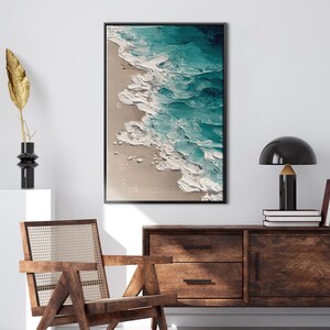 May include: Abstract framed artwork depicting a beach scene with turquoise waves and white foam washing onto a sandy shore. The painting has a textured, three-dimensional appearance, framed in a black border.