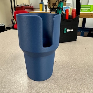 May include: A navy blue cup holder with a textured surface. The cup holder has a unique design with a slot for a handle. The cup holder is sitting on a grey surface.