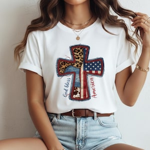 Op de afbeelding: Wit T-shirt met een kruisontwerp met patriottische elementen. Het kruis bevat luipaardprint, denim, de Amerikaanse vlag en koeienhuidpatronen. De woorden "God bless" en "America" staan op het kruis.