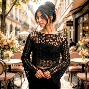 May include: Black crochet top with long sleeves and a unique open weave design. The top is cropped, revealing the midriff. The model is wearing black pants. The background shows a European cafe setting.