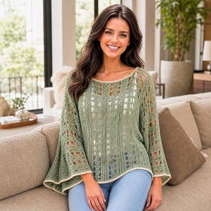 May include: A sage green knit sweater featuring an openwork lace pattern, a wide boat neckline, and flared sleeves. The garment has a light cream trim along the hem and cuffs, paired with light blue denim jeans worn underneath the loose-fitting top.