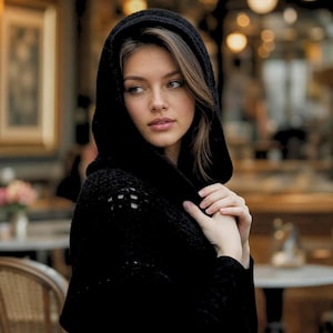 May include: A black hooded garment featuring a textured, open-knit crochet pattern. The hood is pulled up over the head, framing the face with a soft, dark fabric. The material appears lightweight and breathable, with a consistent deep black color.