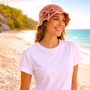 Puede incluir: Un sombrero cloche rosa tejido a ganchillo con un lazo. El sombrero tiene un borde festoneado y un ribete de color beige claro. La persona lleva una camiseta blanca y sonríe en una playa.