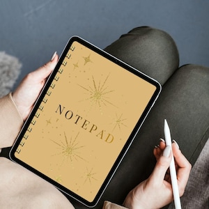May include: A gold-colored notepad with the word "NOTEPAD" in gold lettering. The cover features gold starburst designs. The notepad is spiral-bound and held in a person's lap, with a white stylus nearby.