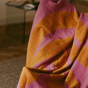 May include: A folded blanket draped over a chair, featuring alternating wide stripes of pink and orange. The blanket has a textured appearance and a fringed edge. The color scheme creates a warm and inviting aesthetic.