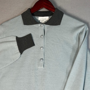May include: A light blue long-sleeve knit shirt featuring a dark gray collar and matching dark gray sleeve cuffs. The shirt has a partial button-down placket with four round light blue buttons and a white brand label reading Schrader.