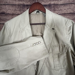 Może przedstawiać: Beżowa marynarka z klapami z wycięciem, naszywanymi kieszeniami i łokciami. Marynarka ma trzy guziki na każdym mankiecie i wisi na drewnianym wieszaku. Wnętrze ma podszewkę z siatki.