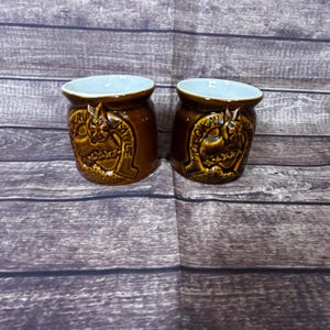 May include: Two brown ceramic mugs with a light blue interior. Each mug features a raised design with the words "Molokai Mule" and a decorative emblem. The mugs are set against a wooden background.