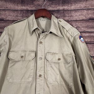 May include: Tan button-up shirt with two chest pockets and shoulder epaulets. The shirt has a classic collar and is hanging on a wooden hanger. A patch with a circular design is visible on the upper sleeve.