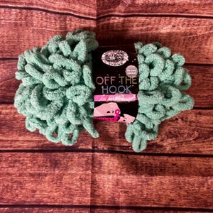 May include: A ball of mint green Off The Hook Glitz yarn, with a label that reads "Off The Hook Glitz" and "No tools needed!" The yarn is a soft, looped texture, perfect for crafting. The background is a wooden surface.