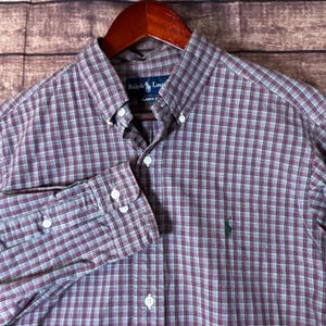 Op de afbeelding: Een button-down shirt met een ruitpatroon in tinten bordeauxrood en lichtblauw. Het shirt heeft een klassieke kraag, lange mouwen en een groen geborduurd logo op de borst. Het shirt hangt aan een houten hanger.