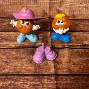 May include: Three vintage Mr. Potato Head toy parts. Two potato-shaped heads with painted eyes, noses, and mouths, one wearing a pink hat and the other with yellow hair. Includes a pair of purple shoes.