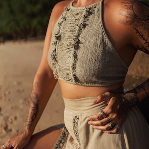 May include: A two-piece outfit featuring a halter top and skirt. The top is a light green, textured knit with fringe detailing. The skirt is a light tan color with a slit and black geometric embroidery along the side. The outfit is suitable for warm weather.
