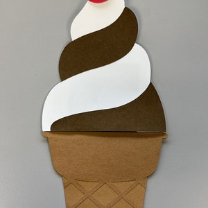 May include: A papercraft ice cream cone with a swirl of white and brown ice cream, topped with a red cherry and green stem. The cone is brown with a diamond pattern.