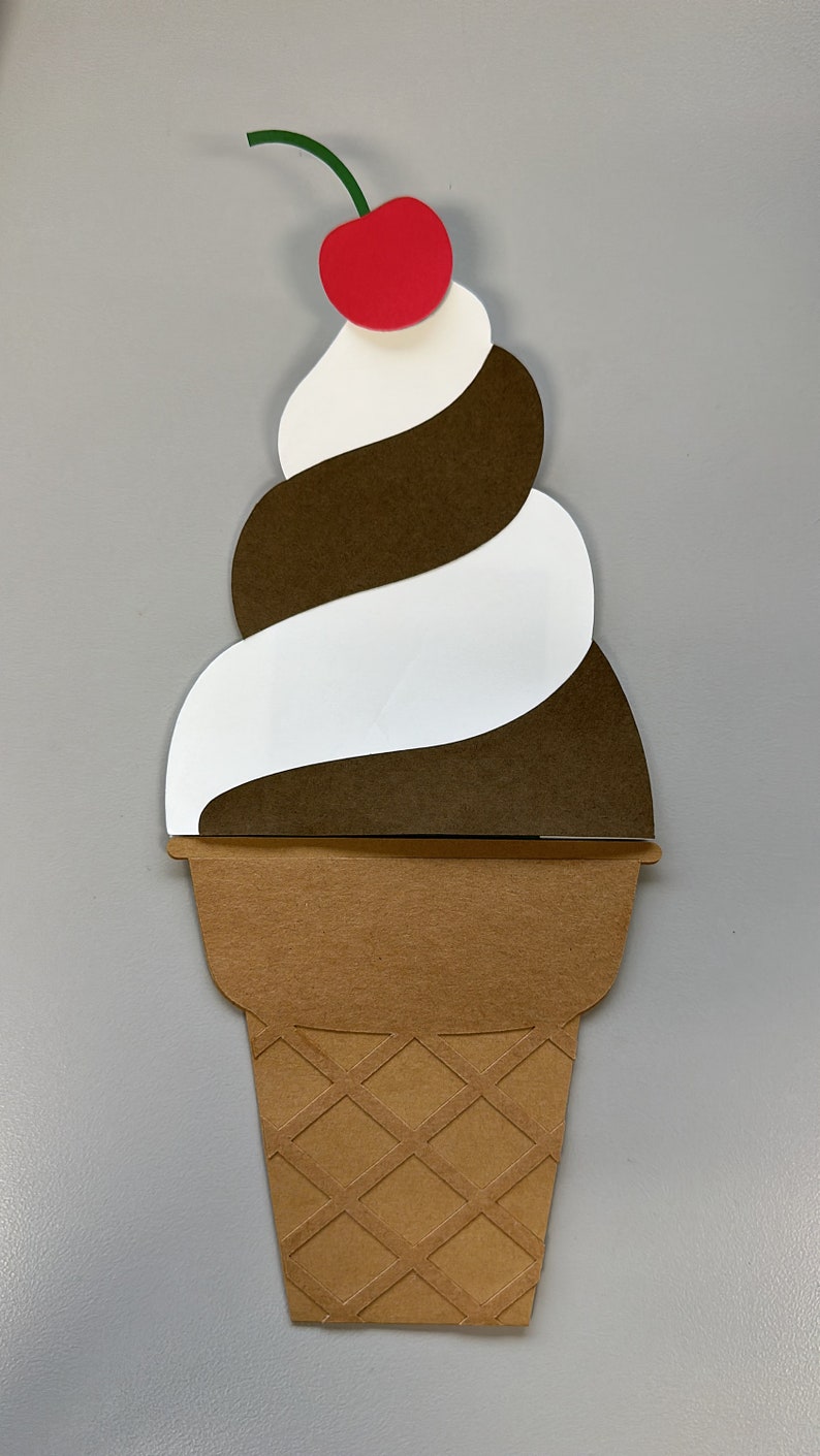 May include: A papercraft ice cream cone with a swirl of white and brown ice cream, topped with a red cherry and green stem. The cone is brown with a diamond pattern.