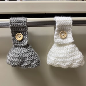 Two crochet towel holders on an oven door. One towel holder is gray and one is white.