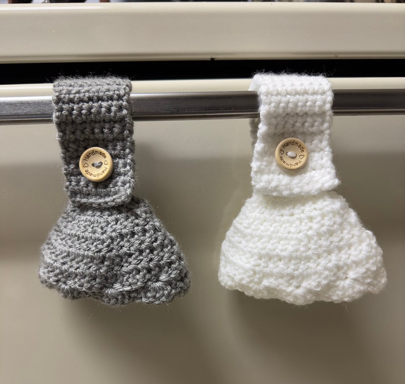 Two crochet towel holders on an oven door. One towel holder is gray and one is white.