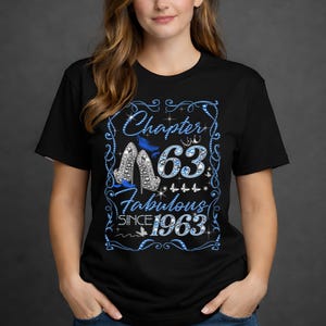 以下が含まれることがあります： 青と銀のデザインが施された黒いTシャツ。デザインには、「Chapter 63 Fabulous Since 1963」の文字と、ハイヒール、蝶、王冠の画像が含まれています。テキストと画像は青で縁取られています。