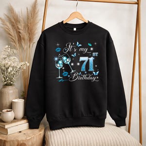 Puede incluir: Sudadera negra con la frase "It's my 71st Birthday". El diseño incluye dos copas de vino, mariposas y el número 71 en azul. El texto es en blanco y azul, ideal para celebrar un 71 cumpleaños.