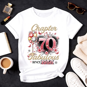 Op de afbeelding: Wit T-shirt met een grafisch ontwerp voor een 70e verjaardag. Het ontwerp bevat de woorden "Chapter 70 Fabulous Since 1956" met een wijnglas, bloemen, vlinders en een kroon. De tekst en graphics zijn in goud, rood en roze.