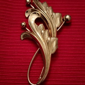 May include: Gold-toned vintage brooch with an ornate, flowing leaf design. The brooch features stylized leaves and small spherical accents. The elegant piece is set against a deep red background, highlighting its intricate details.