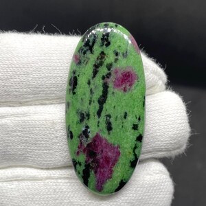May include: An oval-shaped polished gemstone cabochon featuring a vibrant green base colour with prominent deep magenta-pink inclusions and scattered black mineral streaks throughout the smooth, reflective surface of the stone.