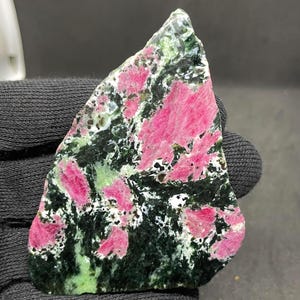 Könnte beinhalten: Eine polierte Platte aus Zoisit-Mineral mit leuchtend magentafarbenen Rubin-Einschlüssen, die in einer dunkelwaldgrünen und schwarzen kristallinen Matrix verteilt sind, in dreieckiger Form mit einer glatten, reflektierenden Oberfläche.