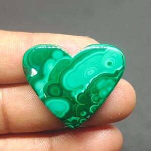 Puede incluir: Una piedra preciosa de malaquita en forma de corazón, que muestra un color verde vibrante con patrones arremolinados. La piedra pulida muestra una mezcla de tonos verdes claros y oscuros, creando un diseño natural único. Esta gema es un bello ejemplo de arte en piedra natural.