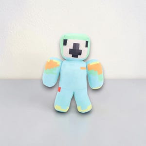 May include: A plush toy in the shape of a blocky figure. The figure has a light blue body, green head, and orange and yellow accents on the arms and legs. The face features a black cross design. The toy is standing on a gray surface.