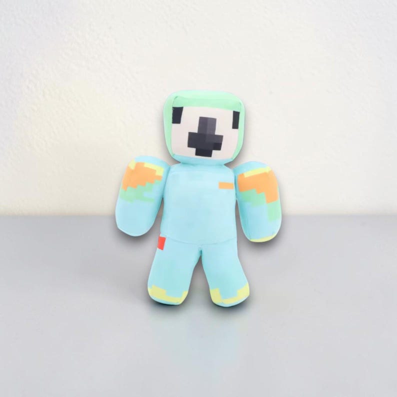 May include: A plush toy in the shape of a blocky figure. The figure has a light blue body, green head, and orange and yellow accents on the arms and legs. The face features a black cross design. The toy is standing on a gray surface.