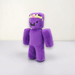 May include: A plush toy in the shape of a purple figure with a pixelated face and a crown. The toy has a blocky design, with arms and legs attached to the main body. The crown is yellow and pink.