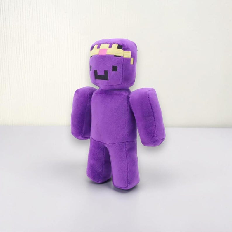 May include: A plush toy in the shape of a purple figure with a pixelated face and a crown. The toy has a blocky design, with arms and legs attached to the main body. The crown is yellow and pink.